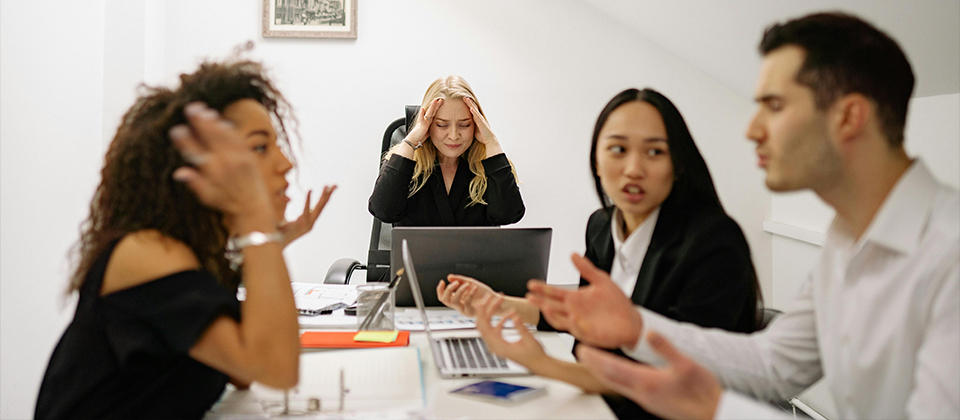 a business woman holds her head in anguish as colleagues argue in the foreground, illustrating the true cost of a cybersecurity incident